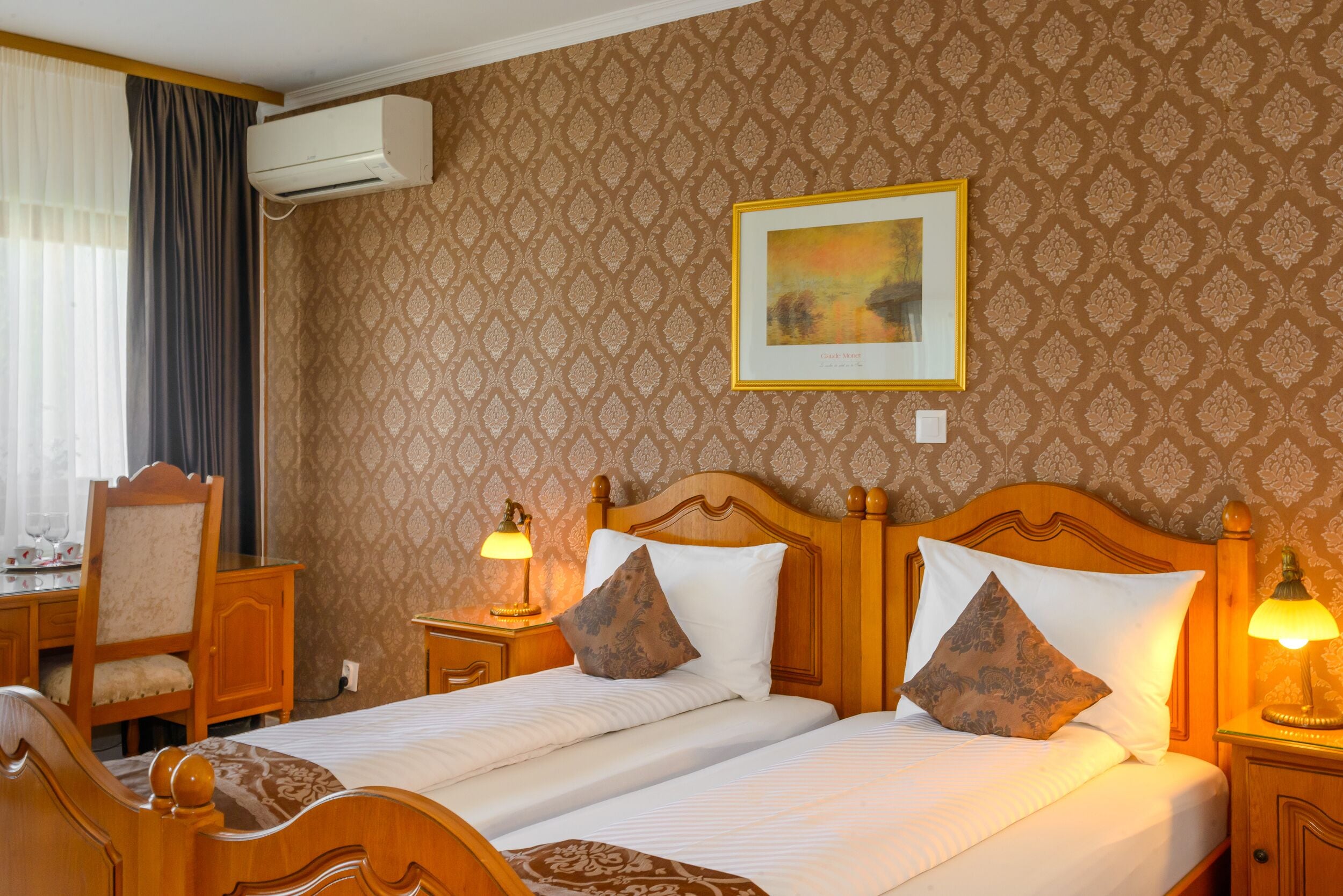 Ein Hotelzimmer mit zwei Betten, einem Holz-Kopfteil, einem Schreibtisch mit Stuhl, einer Lampe und einem gerahmten Bild an der Wand.