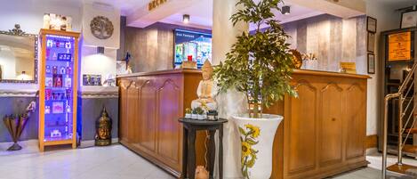 Zona de recepción de un hotel con un mostrador, una planta grande en maceta, una pequeña mesa con una estatua de Buda y una estantería con diversos objetos.