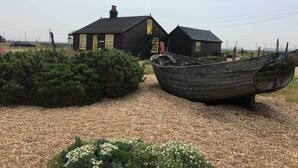 Une maison rustique en bois, avec un toit noir et des portes jaunes, un bateau au premier plan et un chemin de gravier menant à la maison.