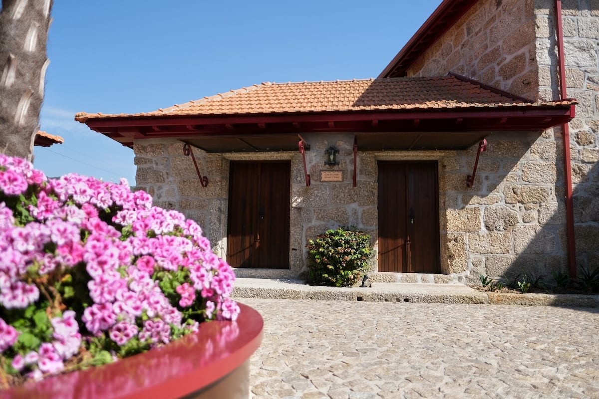 A stone building with a tiled roof, two wooden doors, and a small garden with pink flowers.