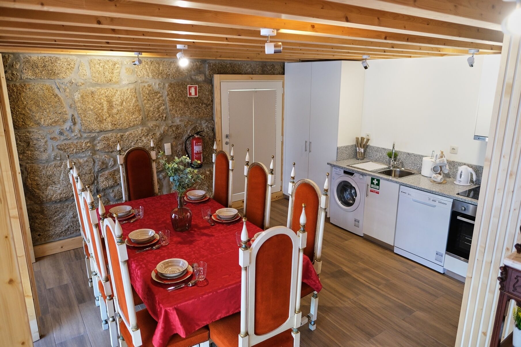 Un coin repas avec une nappe rouge, des chaises en bois et un mur en pierre.