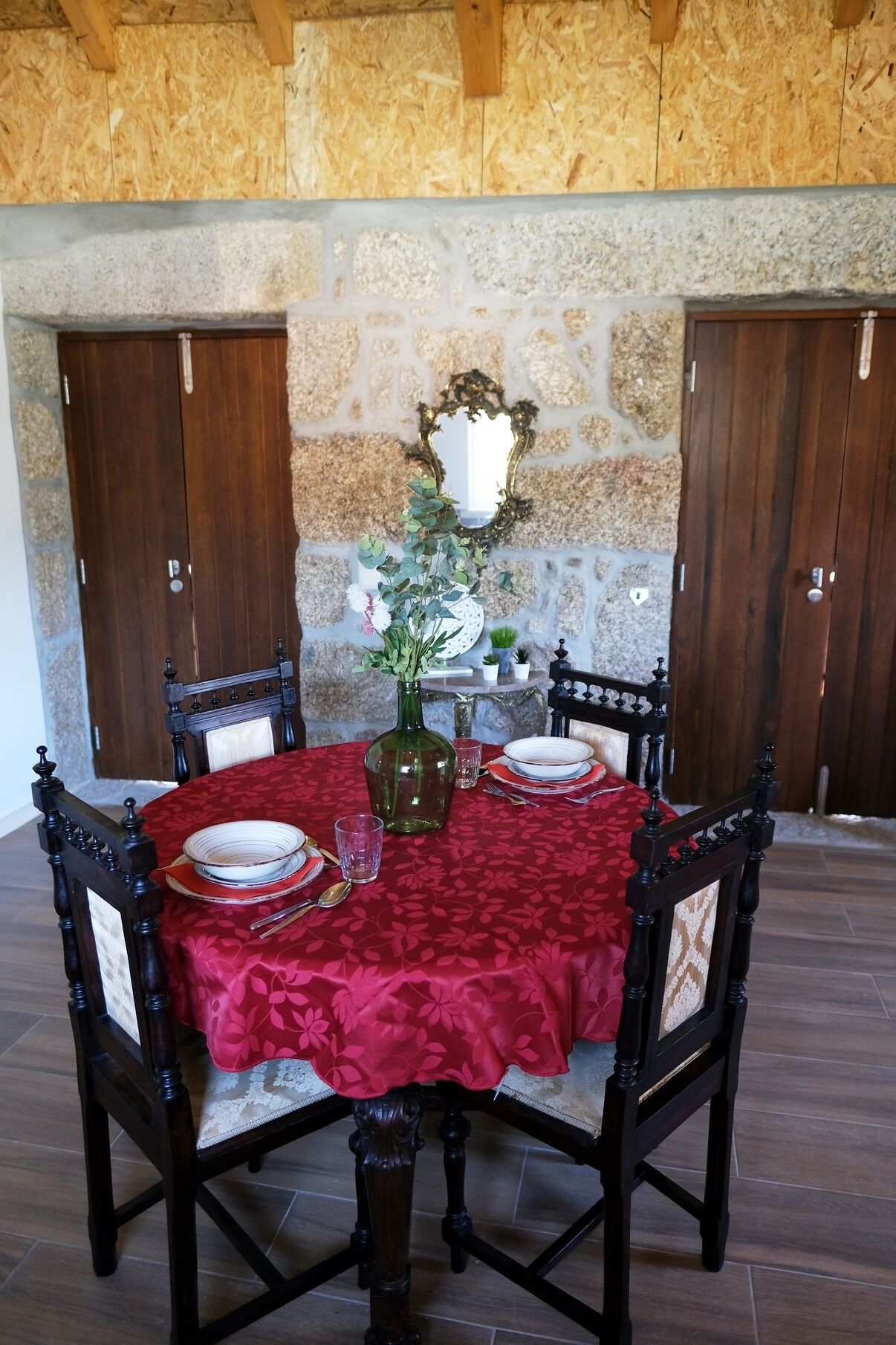 Una tavola da pranzo imbandita per quattro, con una tovaglia rossa, circondata da sedie di legno scuro con sedili imbottiti. Sul tavolo è posata una composizione floreale in un vaso verde.