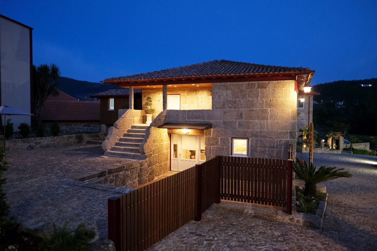 A stone house with a tiled roof, a staircase leading to the entrance, and a gated driveway.