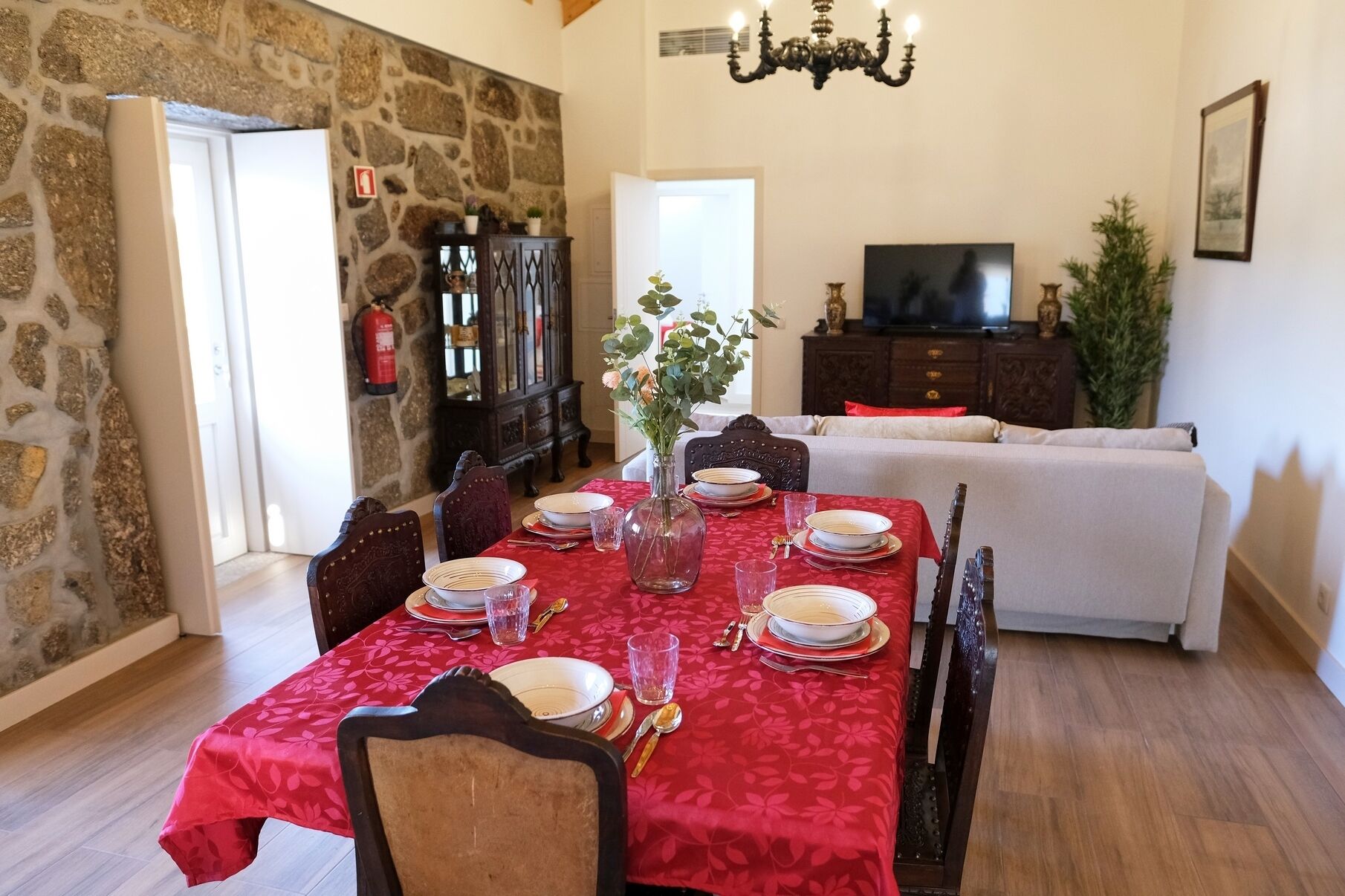 A dining area with a red tablecloth, wooden chairs, and a vase with flowers. A stone wall and a television are visible in the background.