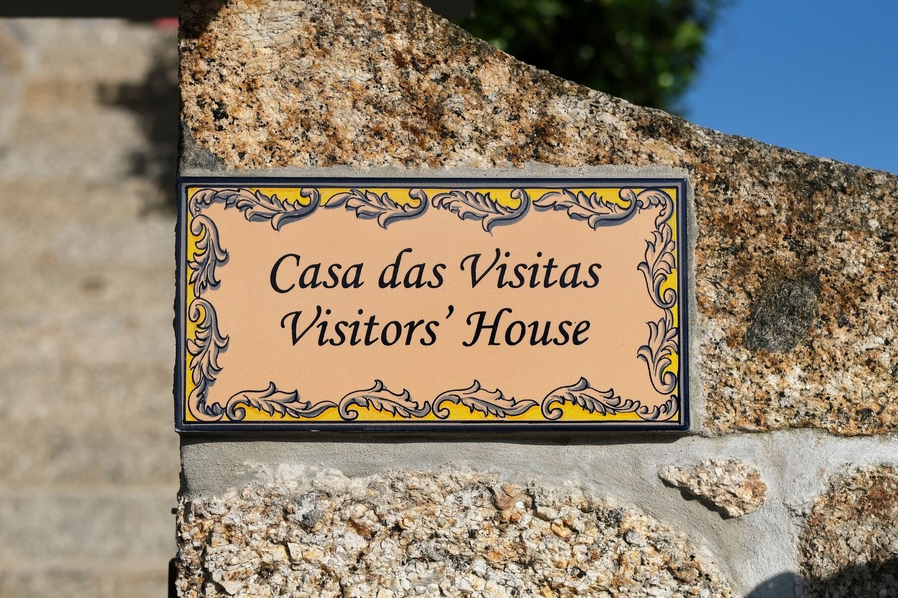 Un letrero con el texto 'Casa das Visitas Visitors' House' sobre una pared de piedra.