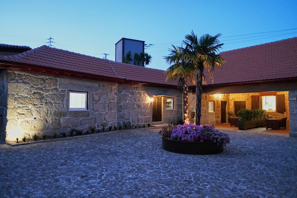 A stone building with a tiled roof, a palm tree, and a flower bed in the courtyard.