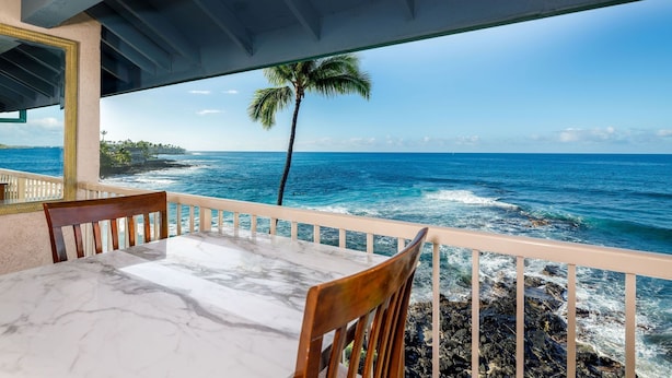 Une table entourée de chaises sur un balcon donnant sur une plage, avec un palmier et les vagues de l’océan en toile de fond.