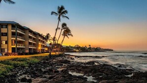 Un hôtel situé en bord de mer, doté de nombreux balcons, de palmiers et offrant une vue sur l’océan au coucher du soleil.