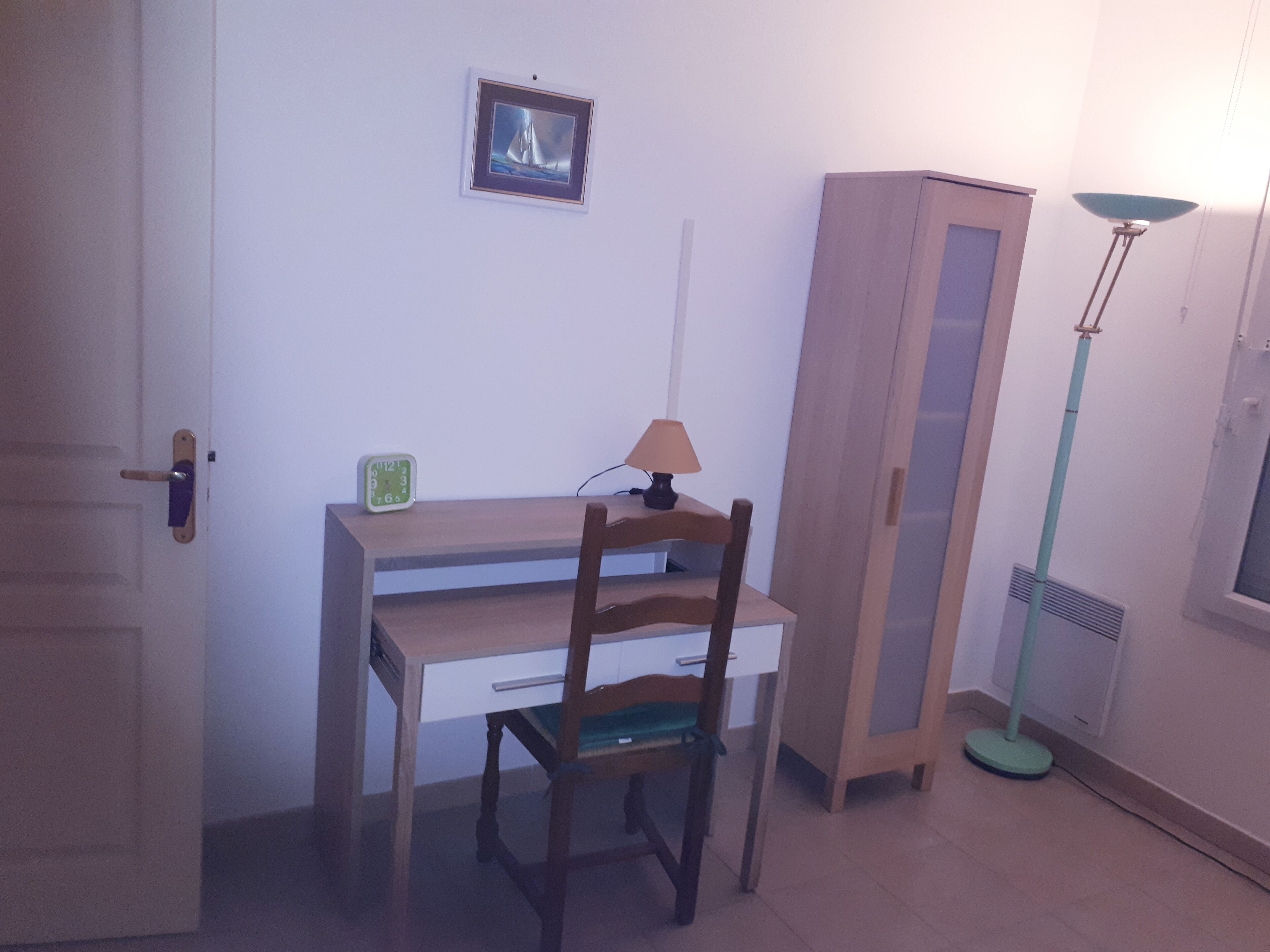 A room with a wooden desk, a chair, a lamp, a wardrobe, and a picture on the wall.