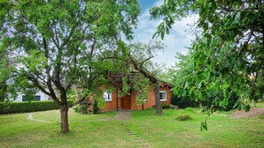 Una casa de ladrillo con puerta y ventanas de madera, rodeada de un jardín cubierto de césped y árboles.