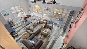 Un vaste espace de vie comprenant un grand canapé, deux petites tables rondes et une table à manger avec des chaises. On y trouve également un ventilateur de plafond, une fenêtre avec des stores et un élément décoratif mural.