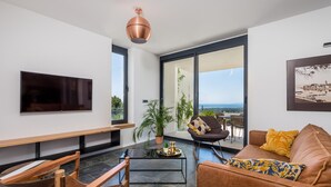 A modern living room with a flat-screen TV, a wooden bench, a sofa with patterned cushions, a coffee table, and a large window offering a view of the sea. - Beach front home in Palit with sauna (Palit)