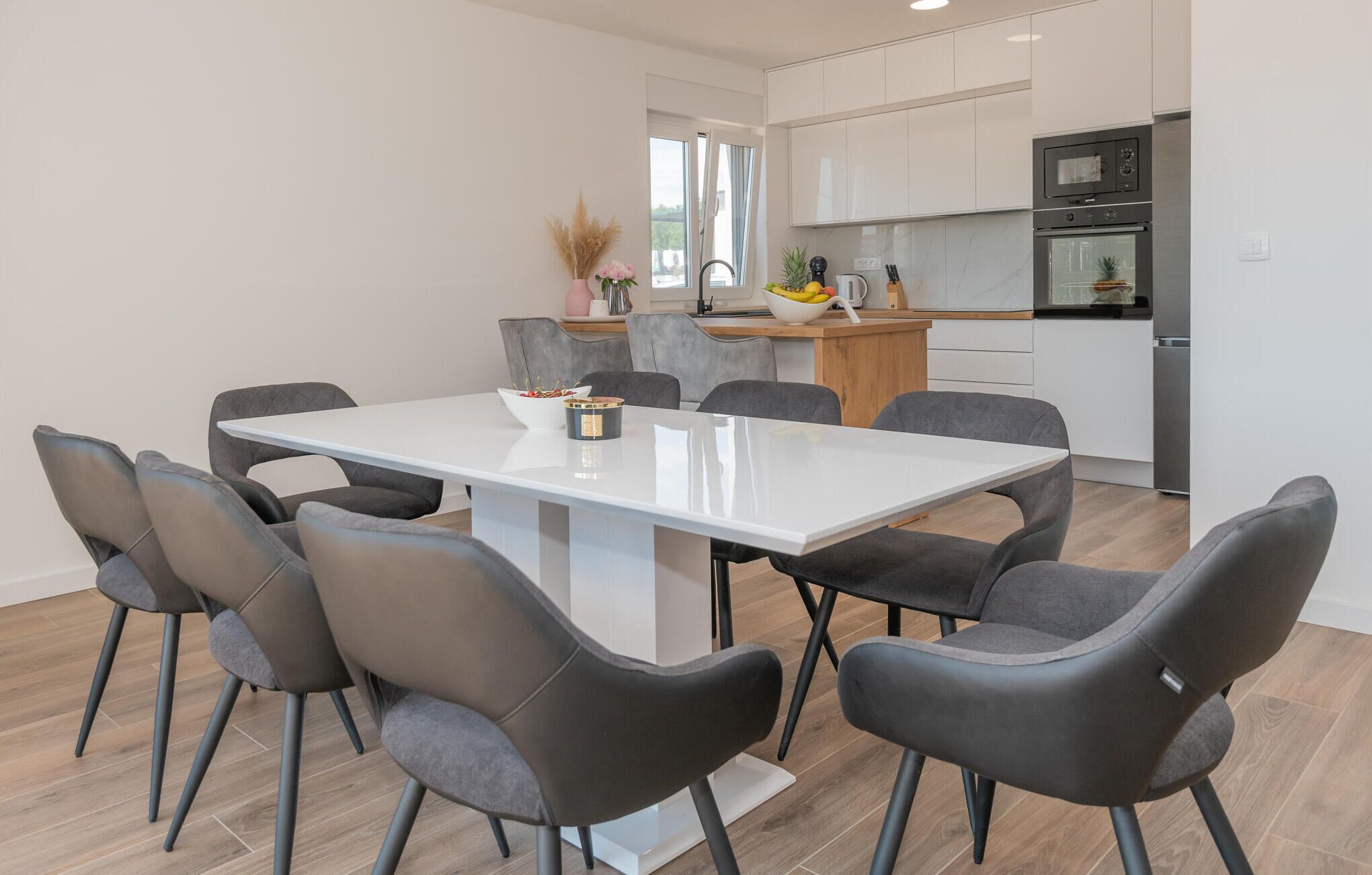 Eine moderne Küche mit weißen Schränken, einem eingebauten Ofen und einem Essbereich mit einem weißen Tisch und grauen Stühlen.