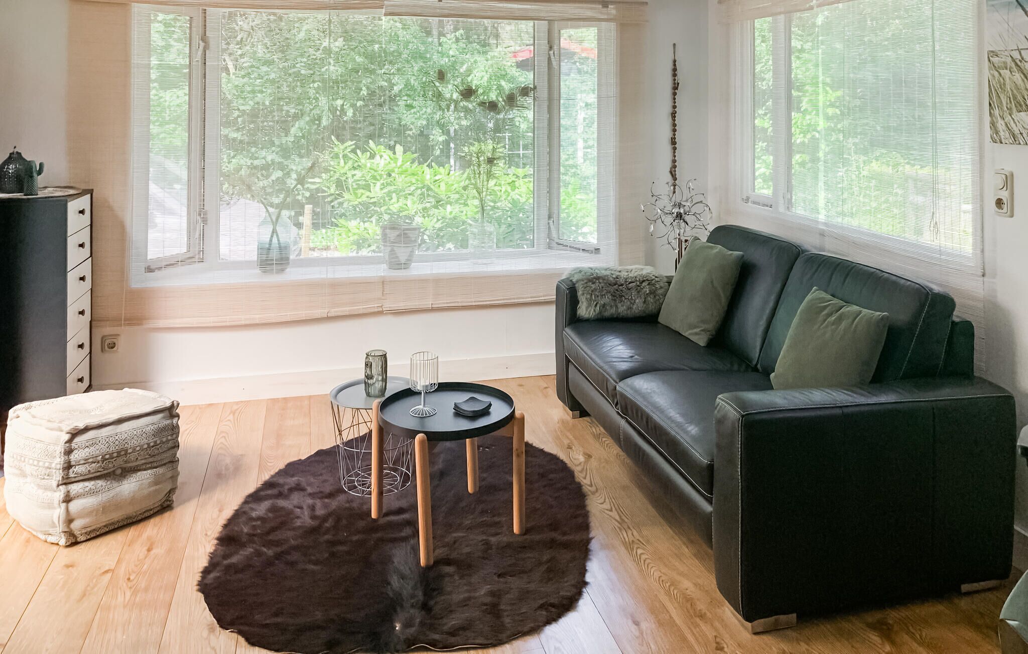 A modern living room with a black leather sofa, a round coffee table, and a large window with a view of greenery.