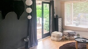 A room with a sliding glass door leading to a garden, a dark wall with a hanging light fixture, a wooden floor, a small round table with a chair, and a dark rug.