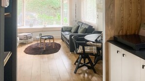 A modern living room with a sofa, a coffee table, and a dining area with chairs. There are large windows providing a view of greenery outside.