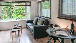 A modern living room with a leather sofa, a dining table set for two, and a view of lush greenery through large windows.
