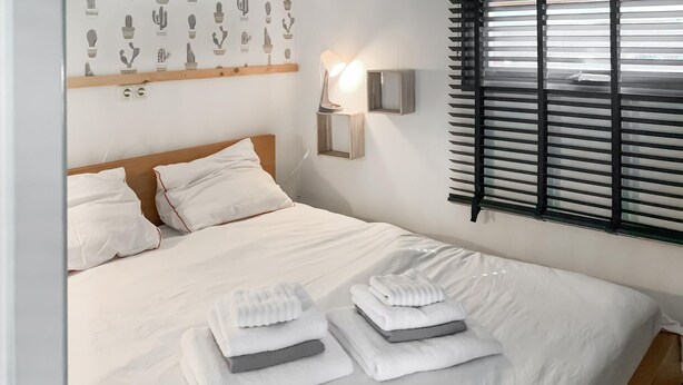 A neatly made bed with white linens and folded towels, a bedside lamp, and a window with blinds.
