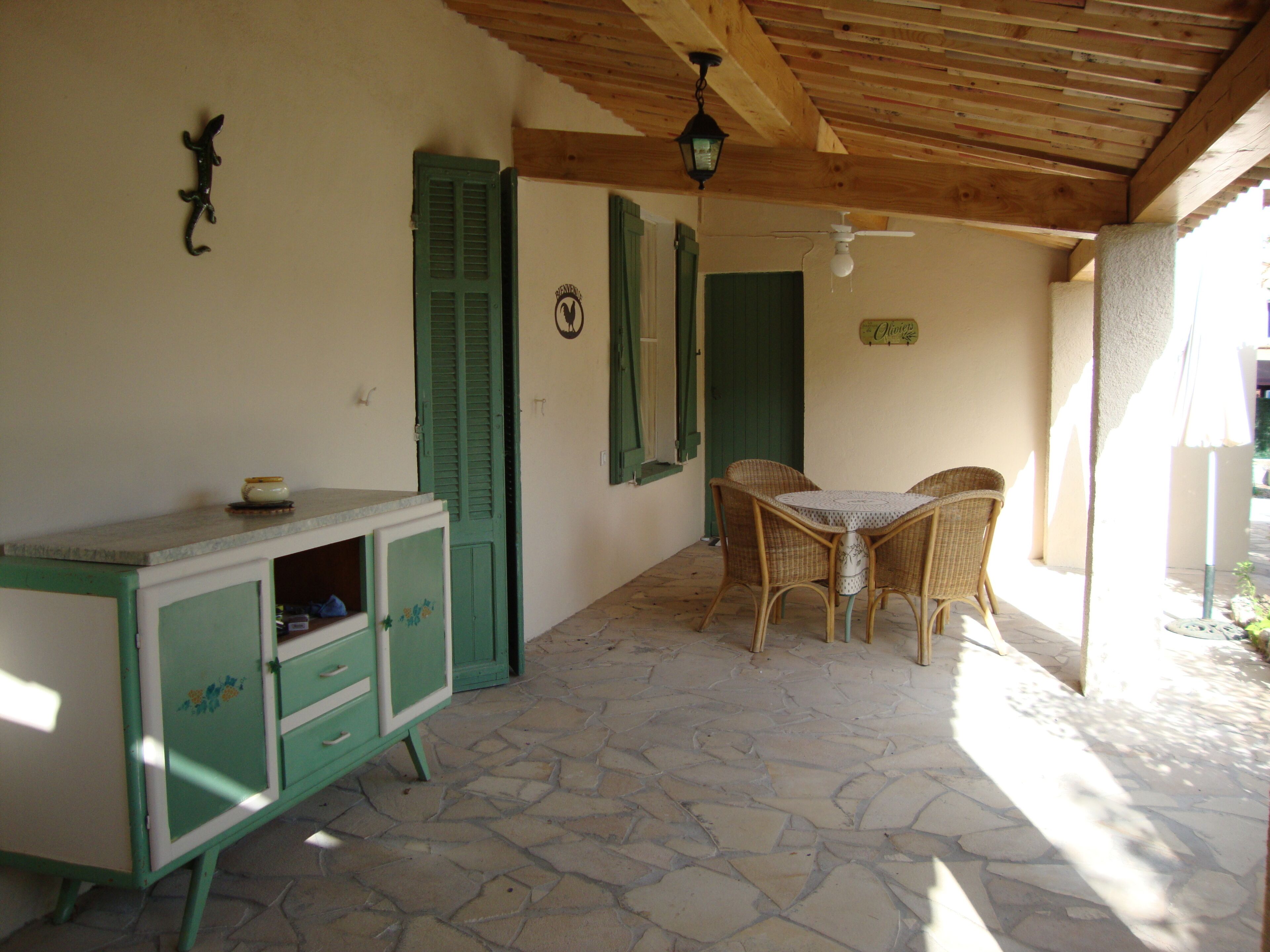 A covered patio area with a stone floor, a green cabinet with drawers, wicker chairs, and a table.