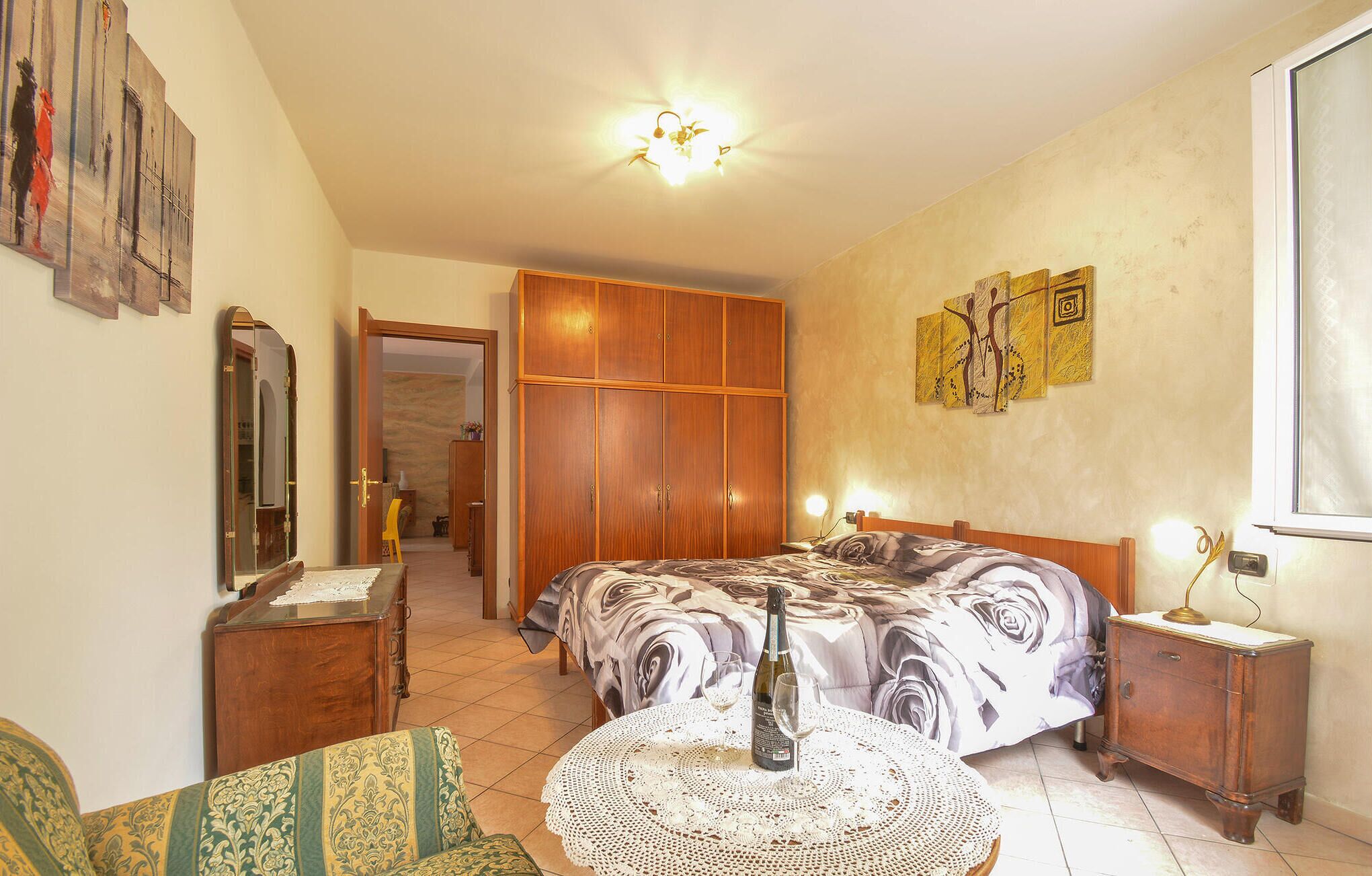 Un dormitorio con cama, mesita de noche, silla, armario de madera, espejo y puerta que da a otra habitación.