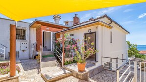 Ein kleines, zweistöckiges Gebäude mit weißer Fassade und Eingang aus roten Ziegelsteinen. Es besitzt einen kleinen Balkon mit Metallgeländer und einem Blumentopf mit rosa Blüten. Das Gebäude befindet sich auf einer gepflasterten Terrasse, wo ein gelber Sonnenschirm Schatten spendet.