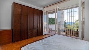Un dormitorio con un armario de madera, una cama y una puerta corrediza de vidrio que da a un balcón con vistas a zonas verdes y montañas.