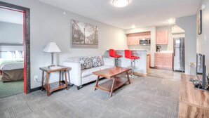 A compact living space with a sofa, a coffee table, a lamp, a painting, a kitchenette, and a TV.