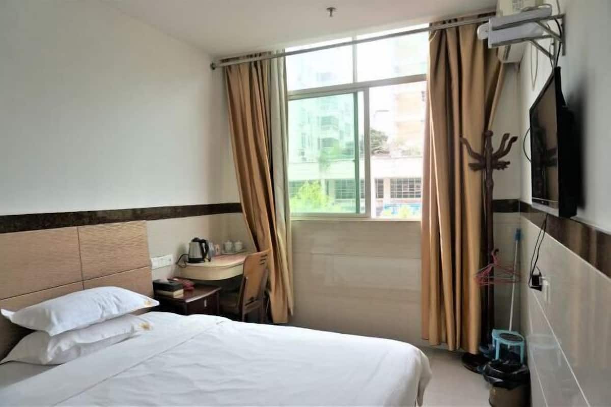 A hotel room with a bed, a small table with a kettle, a TV, and a window with curtains.