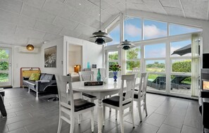 A modern dining area with a white table and chairs, a black sofa, and a large window offering a view of greenery.