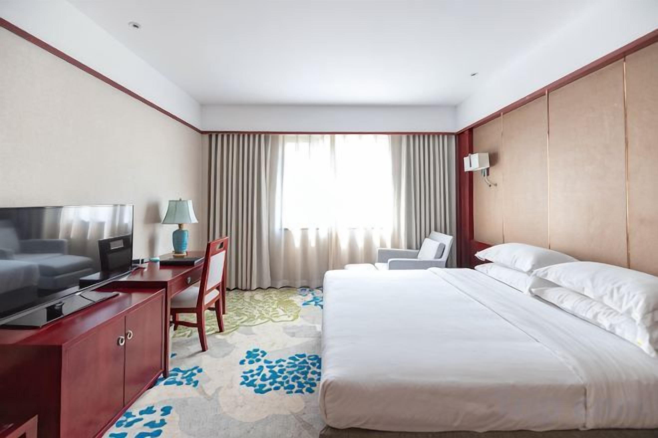 A hotel room with a bed, a desk with a television, a chair, and a window with curtains.
