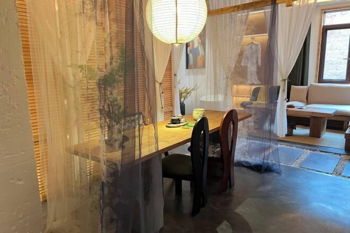 Ein Essbereich mit Holztisch, Stühlen, einer Hängeleuchte und Blick auf einen Wohnraum mit Sofa und Fenster.