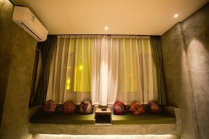 A room with a window covered by sheer curtains, a small table with decorative items, and an air conditioning unit on the wall.