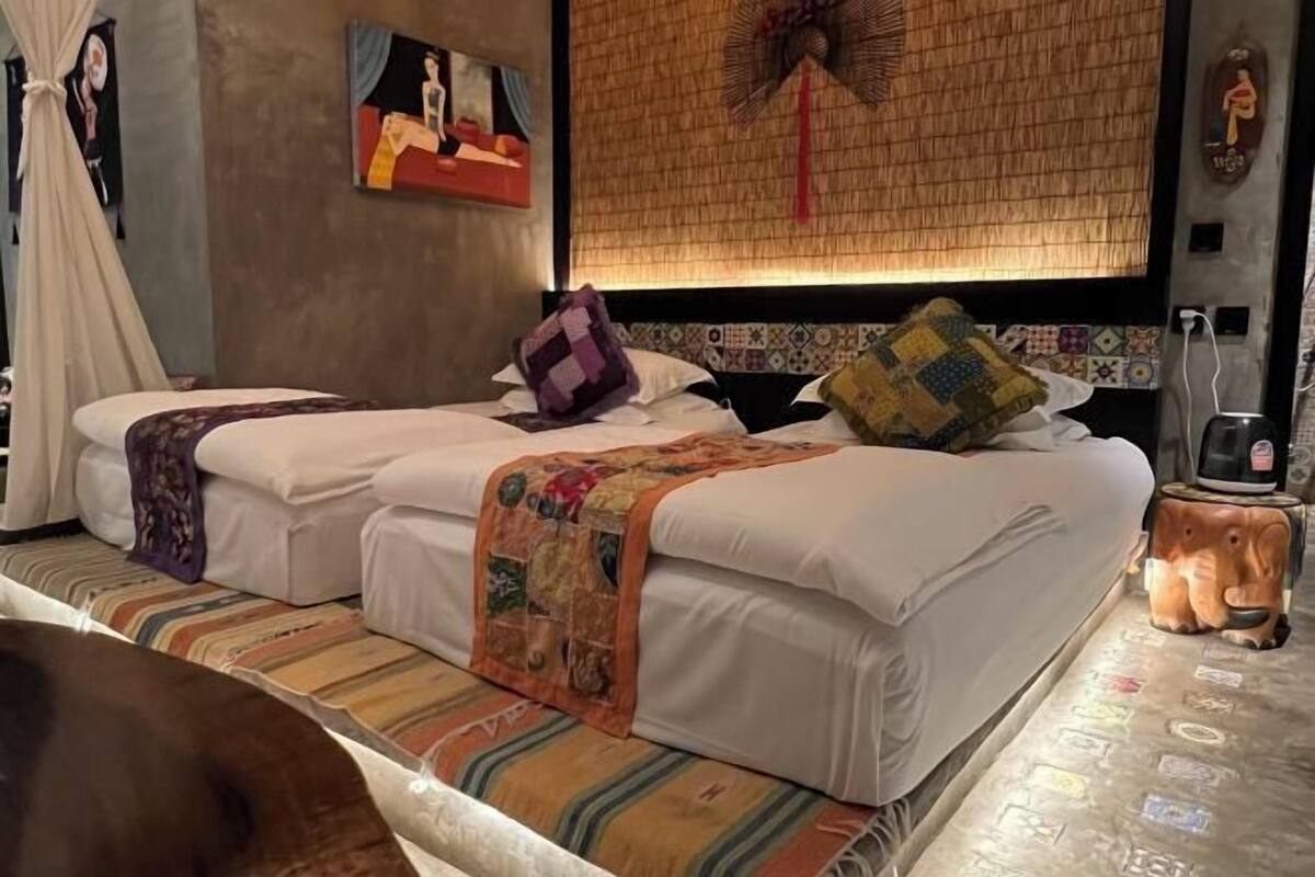 Ein Doppelbett mit zwei Kissen, ein Nachttisch, ein Wandventilator und ein dekorativer Gegenstand auf dem Boden.