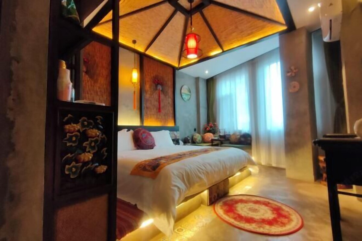 Una habitación de hotel con una cama, cabecero de madera, alfombra estampada en rojo y blanco y un ventilador de techo.