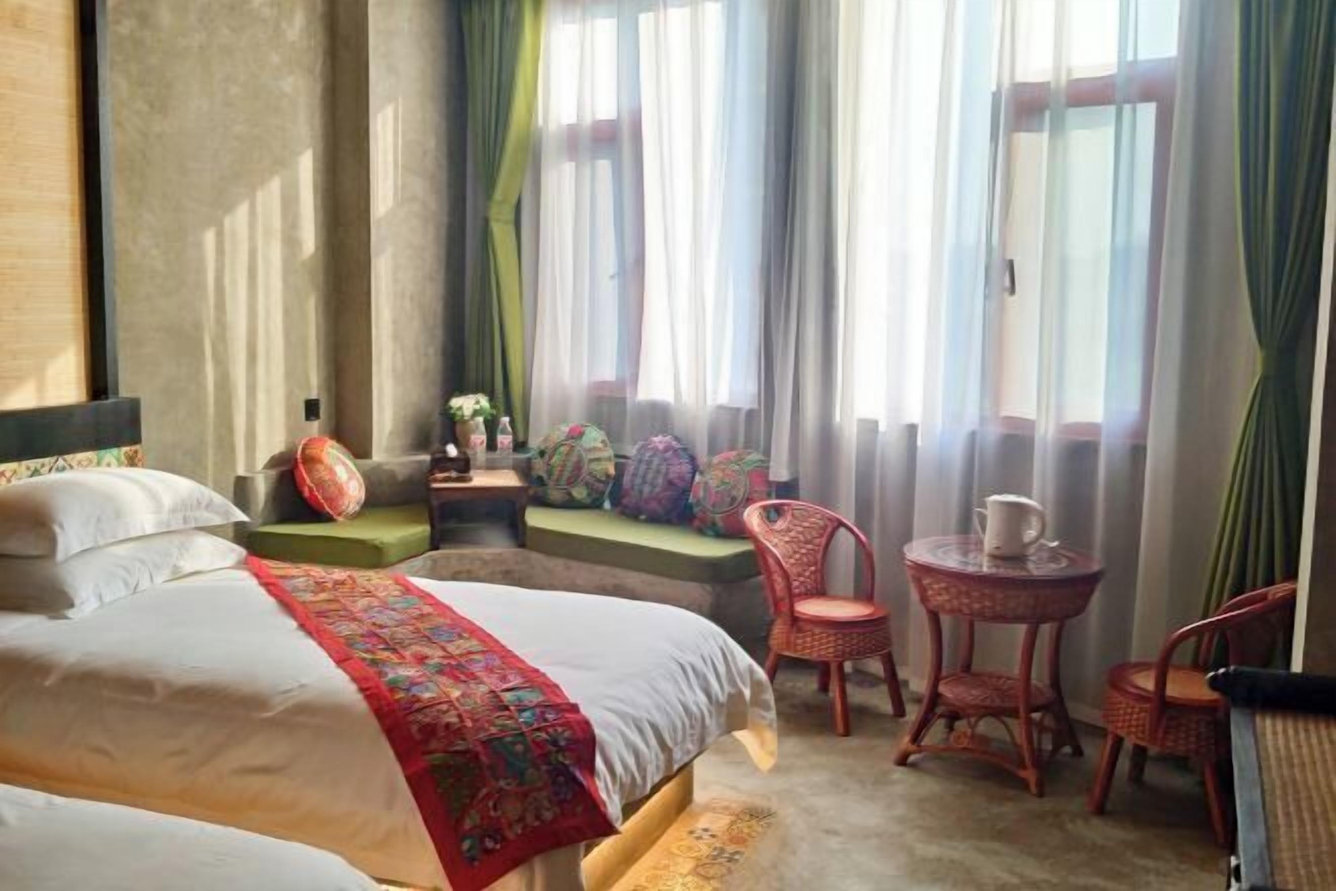 A hotel room with a bed, a small table, two chairs, a bench, and a window with curtains.