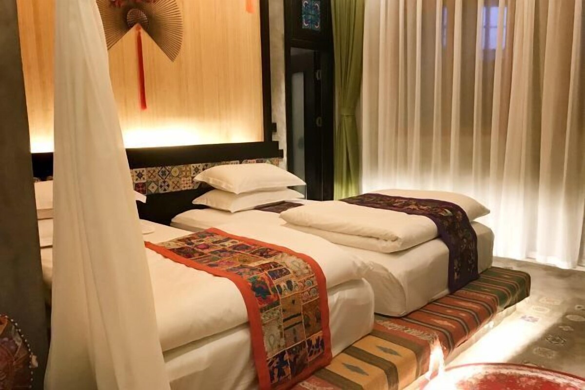 Habitación de hotel con dos camas, cada una con un edredón estampado, una mesita de noche y un ventilador de pared.