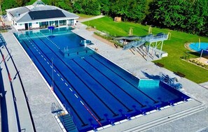 Una gran piscina con múltiples carriles, una zona de estar techada y un pequeño edificio con tejado inclinado.