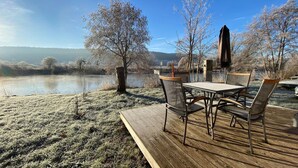 En træterrasse med et bord og stole med udsigt over en sø, omgivet af frostklædte træer og en klar himmel.