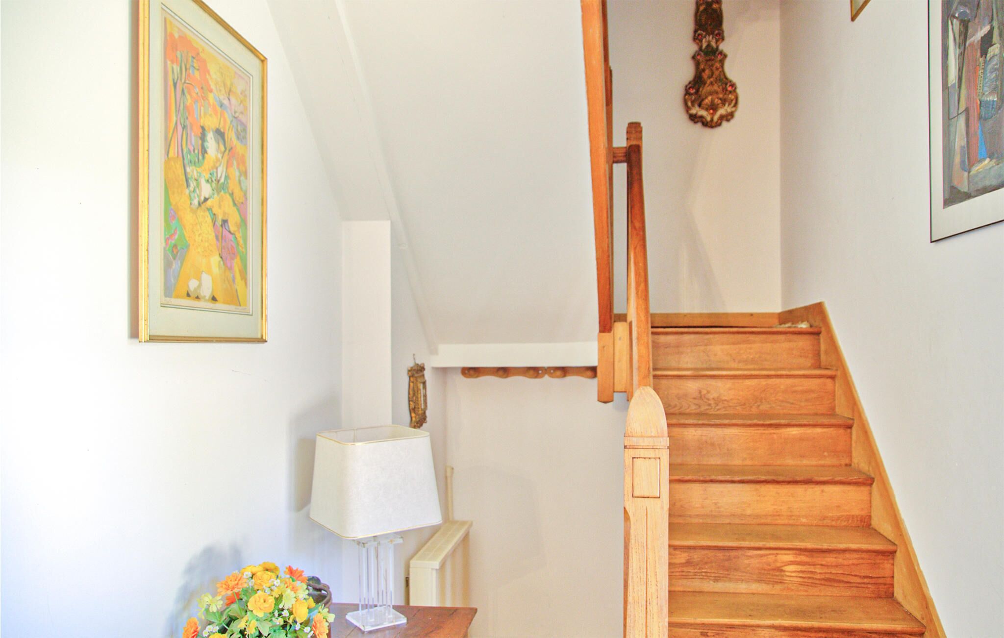 A staircase with wooden steps and a handrail, a white wall with a framed painting, and a table with a lamp and a vase of flowers.