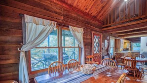 A wooden cabin interior with a large dining table, wooden chairs, and a kitchen area.