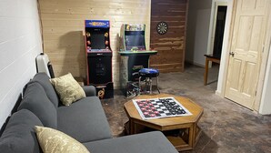 A basement room with a couch, two arcade machines, a dartboard, and a chess table.