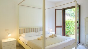 Ein Himmelbett mit weißer Bettwäsche, ein Nachttisch mit Lampe und Blick ins Grüne durch eine offene Tür.