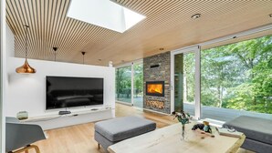 A modern living room with a large TV, a fireplace, and large windows overlooking a forest. - Gorgeous home in Bryrup with kitchen (Bryrup)