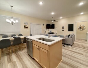 Enjoy cooking and relaxing in this spacious kitchen and living area, featuring a central island with sink, dishwasher, and countertop, plus a comfortable sofa and wall-mounted TV.