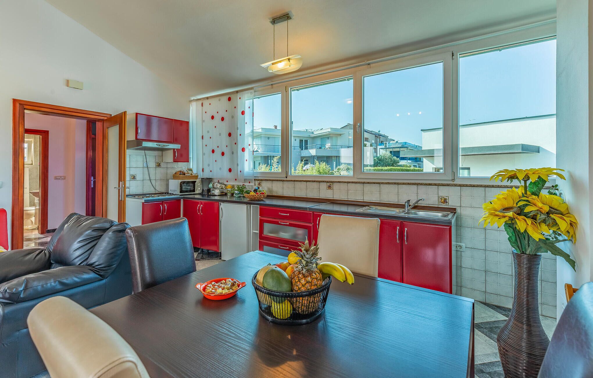 Une cuisine moderne avec des meubles de rangement rouges, un coin repas avec une table et des chaises, et de grandes fenêtres offrant une vue sur le quartier.