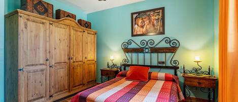 A bedroom with a large wooden wardrobe, a bed with a colorful quilt, a painting on the wall, and two bedside tables with lamps.