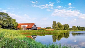 Ein traditionelles niederländisches Haus mit rotem Dach, gelegen an einem ruhigen See mit üppiger Vegetation und Bäumen im Hintergrund.
