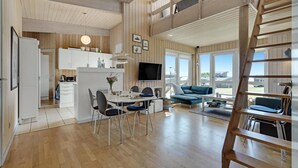 A spacious living area with a dining table, chairs, a sofa, and a TV. There is a kitchen area with cabinets and a refrigerator. A wooden ladder is visible in the corner.
