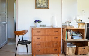 Une commode en bois avec des tiroirs, une chaise en osier, une petite table avec un vase et des fleurs, et une bibliothèque remplie de livres et d’objets décoratifs.