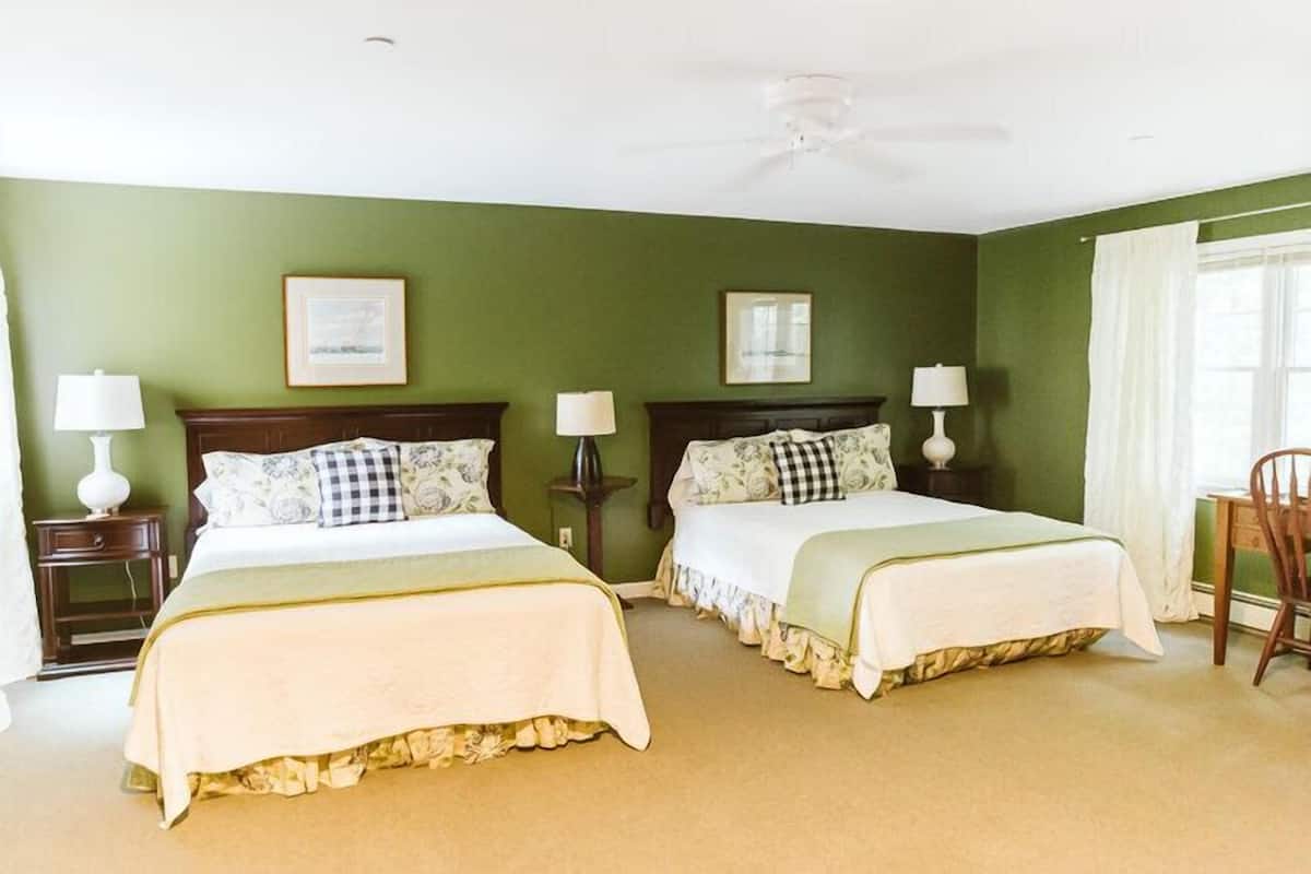 A room with two beds, each with a white bedspread and matching pillows, a ceiling fan, and a window with white curtains.